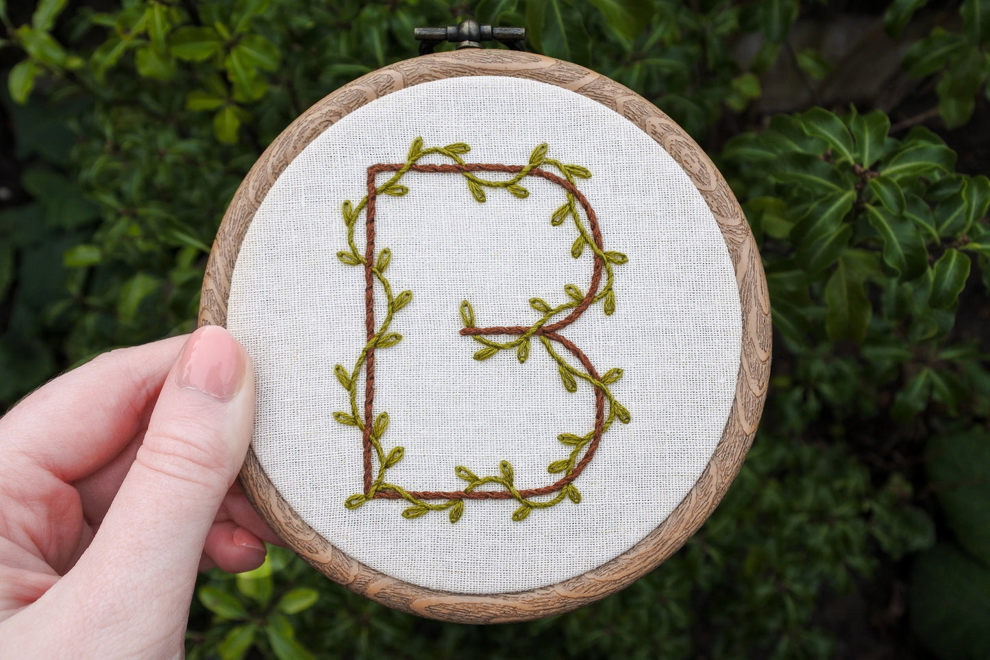 Hand holding hand embroidered botanical letter B hoop against out of focus dark green leaf background.