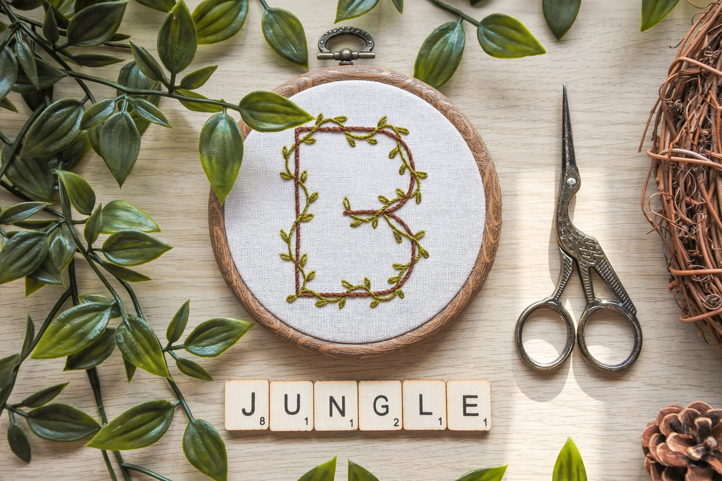 Botanical letter 'B' embroidery hoop displayed on neutral fabric with wooden Scrabble letters spelling "Jungle" underneath.