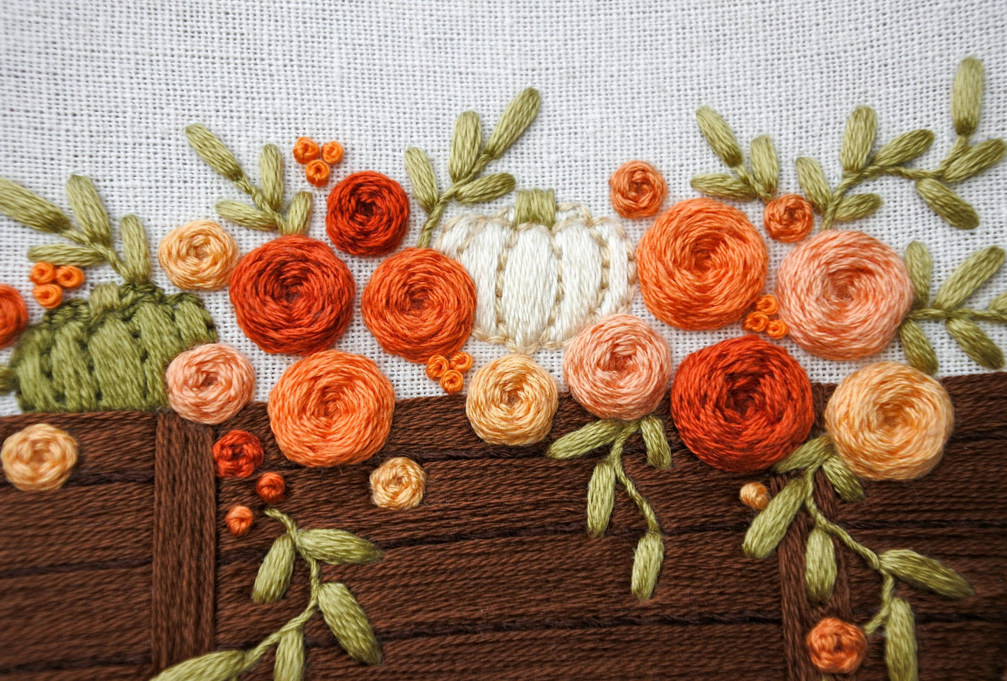 Closeup of hand embroidered orange, red and peach roses with white and green pumpkins in a brown wagon with trailing green foliage. Stitched on light neutral cotton fabric.