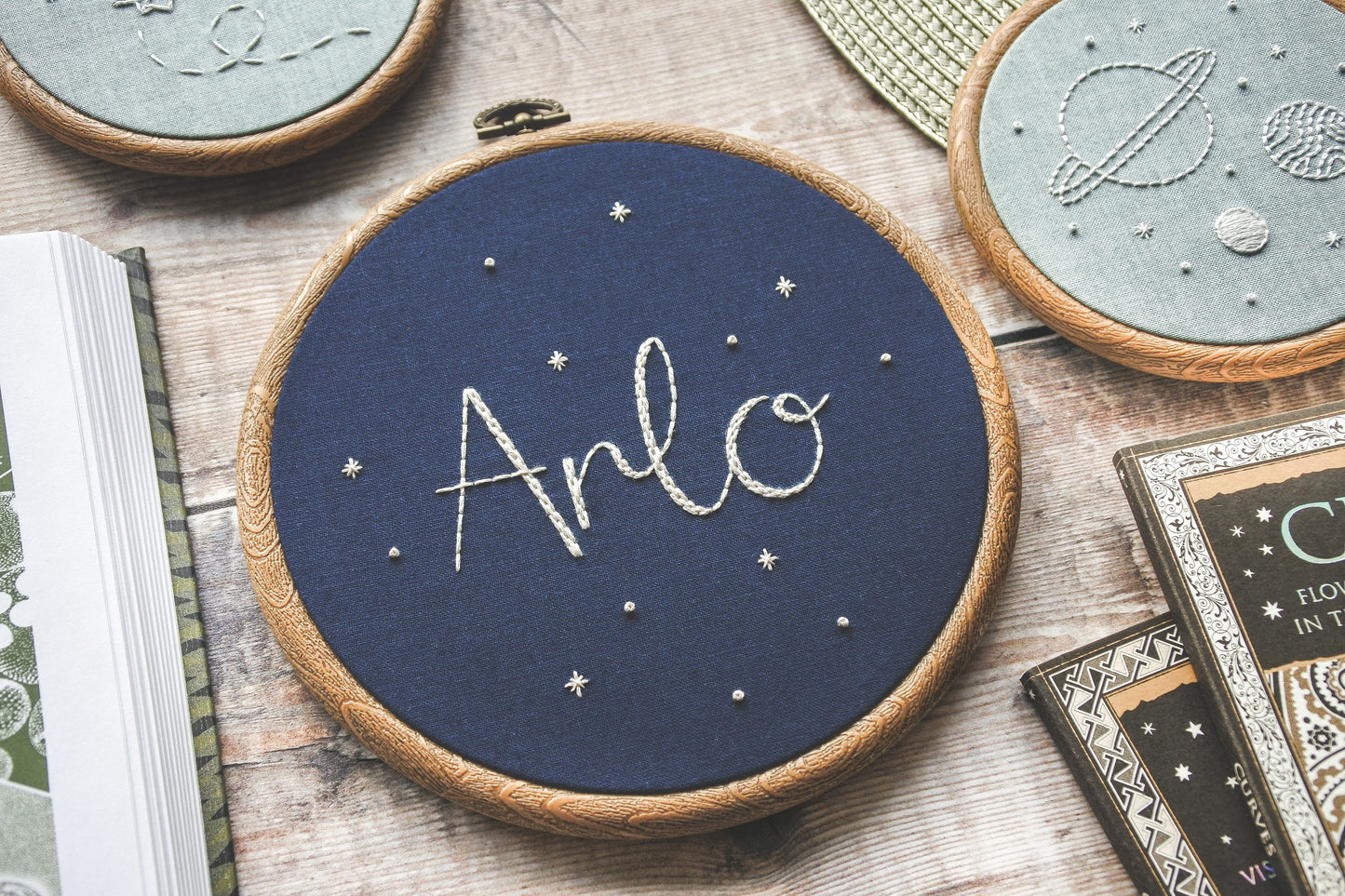 Hand embroidered name hoop with the name Arlo stitched in white thread on navy fabric, framed in a 6-inch wood-effect hoop
