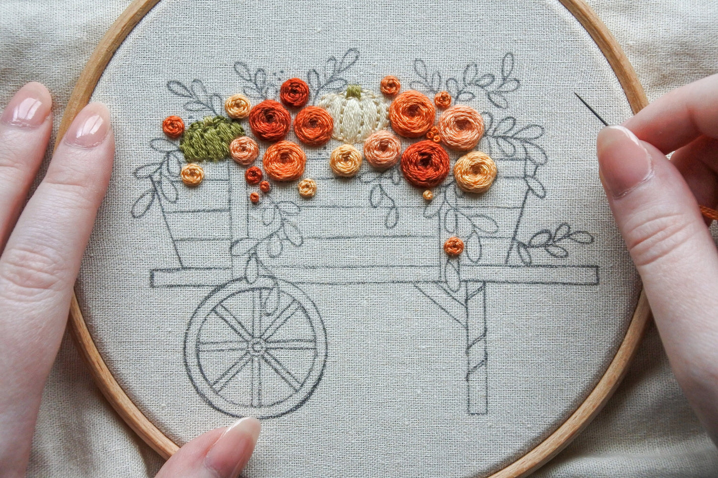 Hand holding a needle and thread while stitching flowers and pumpkins on an autumn wagon embroidery pattern