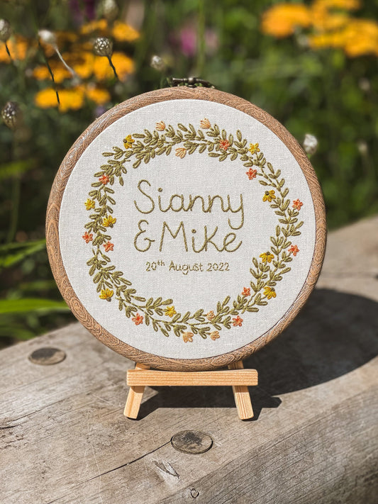 Embroidered hoop with “Sianny and Mike” and their date, shown on a wooden easel outdoors with warm summer tones