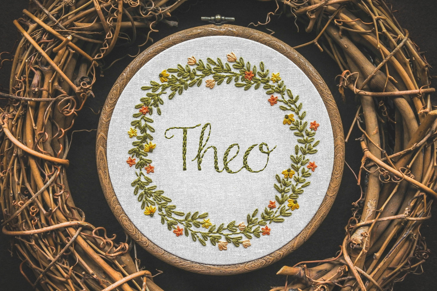 Hand embroidered summer wreath name hoop with the name Theo stitched in the centre, featuring bright floral details and warm green leaves.
