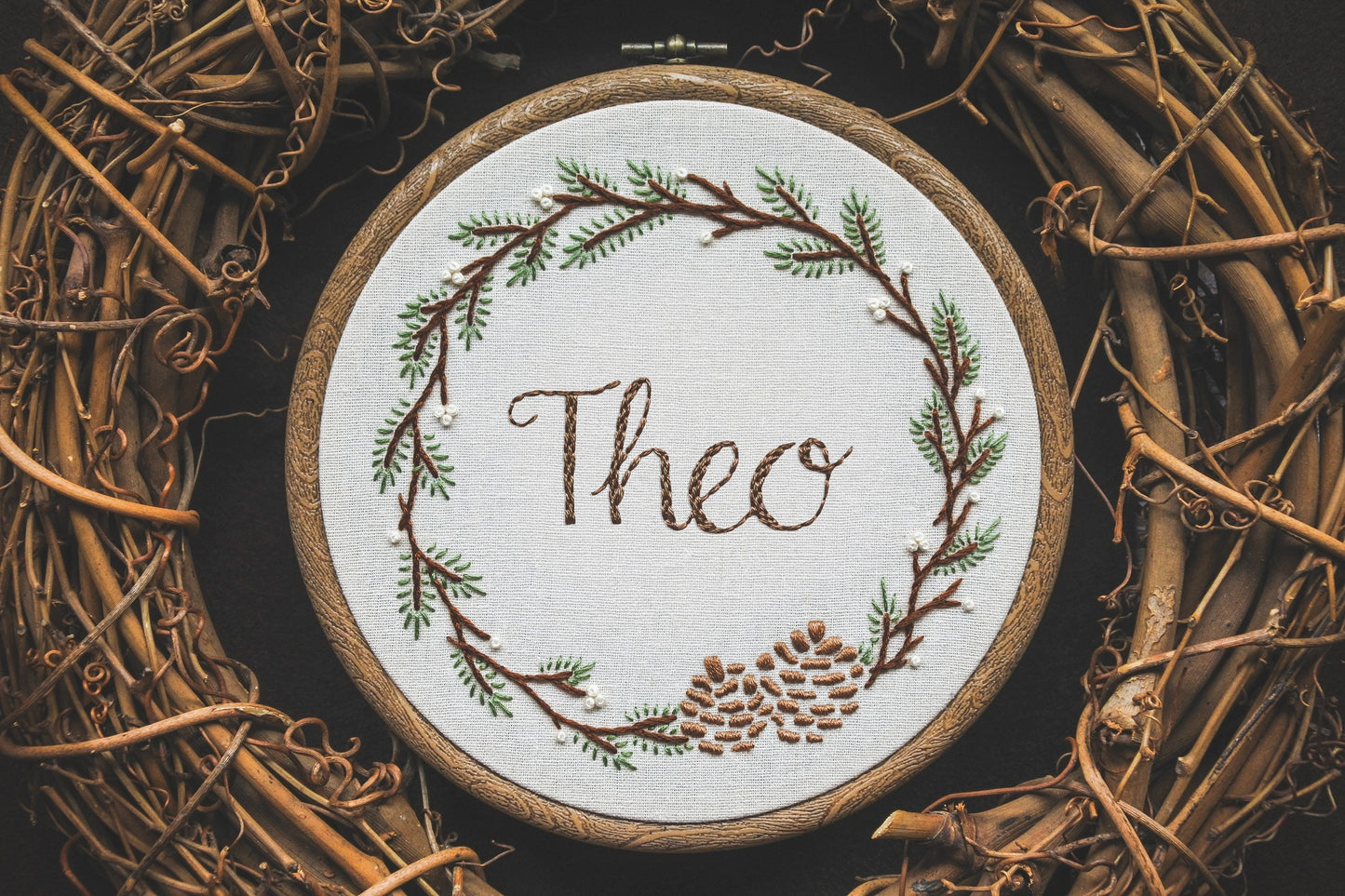 Hand embroidered winter wreath name hoop with 'Theo' stitched in green thread, surrounded by pinecones and evergreen foliage