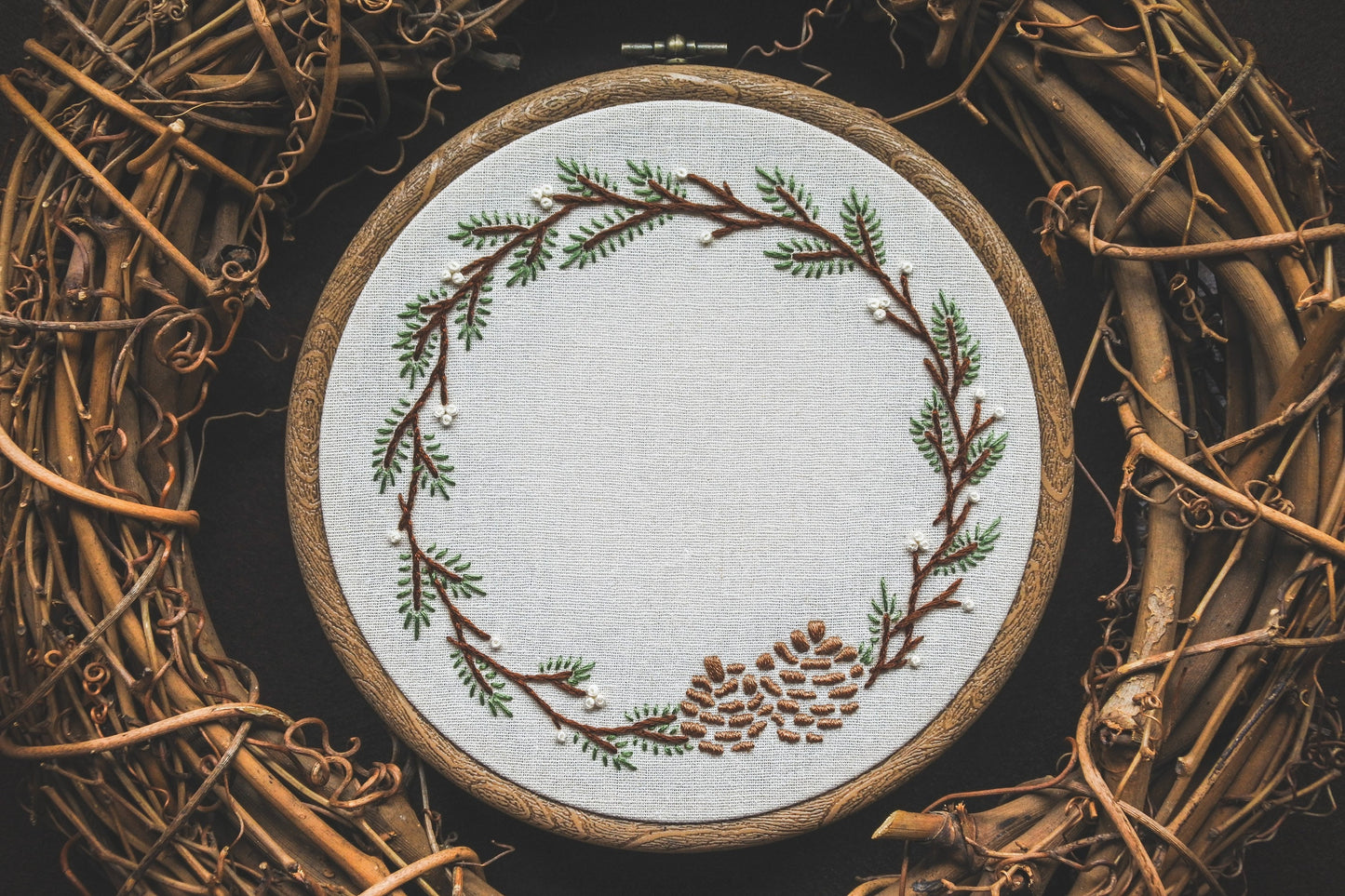 Winter wreath embroidery with pine branches and pinecones, blank in the centre ready to be customised with a name. The embroidery is in a wood effect hoop frame.