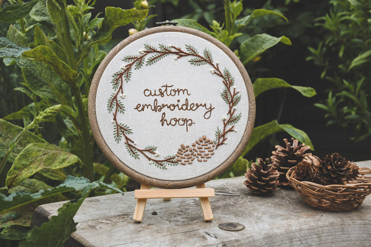 Winter wreath embroidery hoop displayed outside on a mini wooden easel, surrounded by greenery and pinecones.