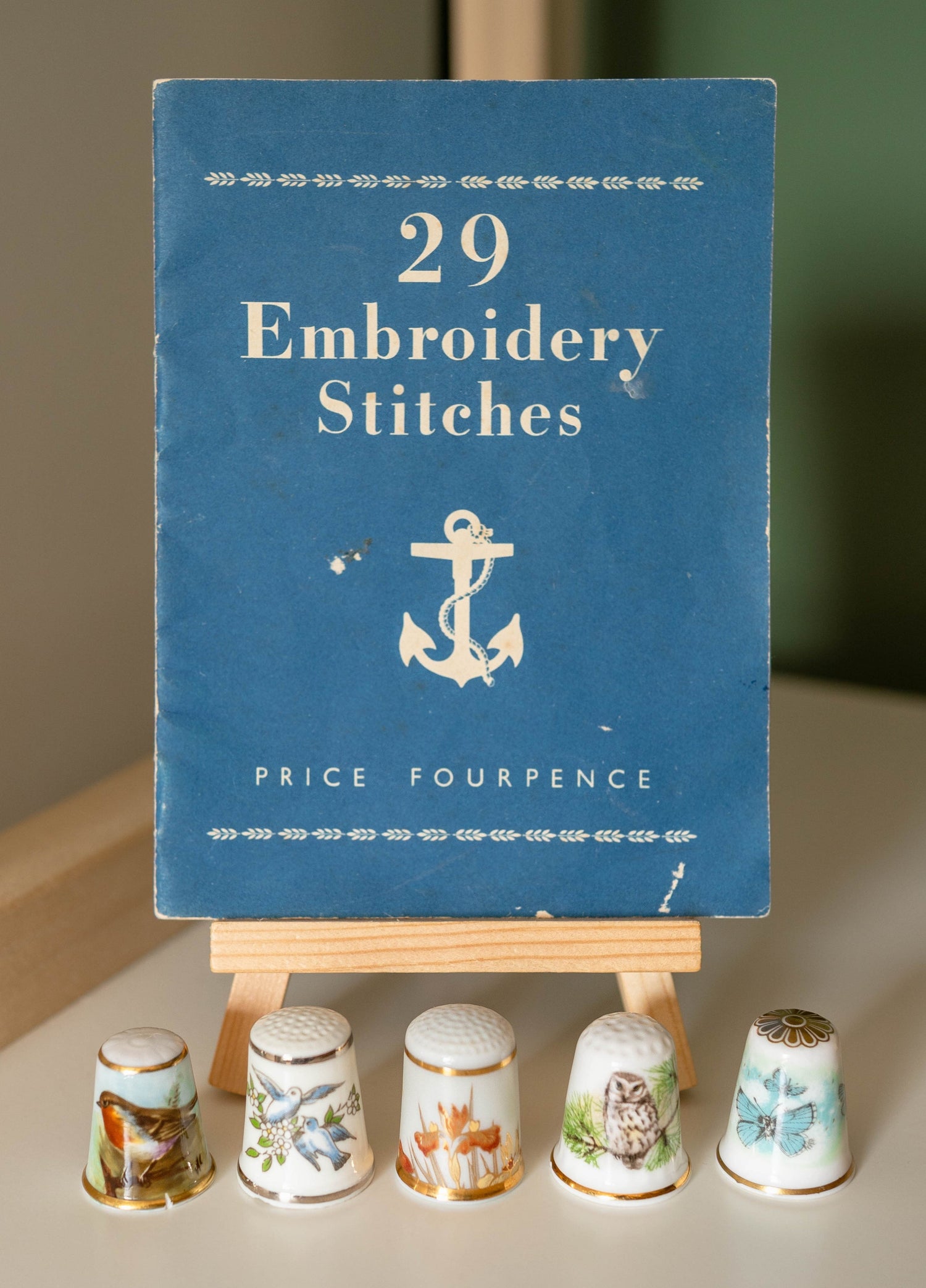 Small blue book of 29 embroidery stitches displayed on a mini wooden easel with five decorative thimbles lined up in front of it.