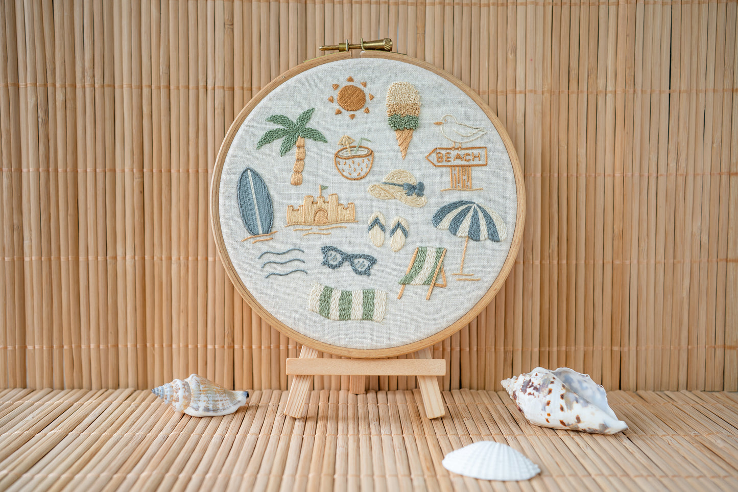 Embroidery hoop with beach-themed designs stood on a mini easel on a wooden surface with shells.