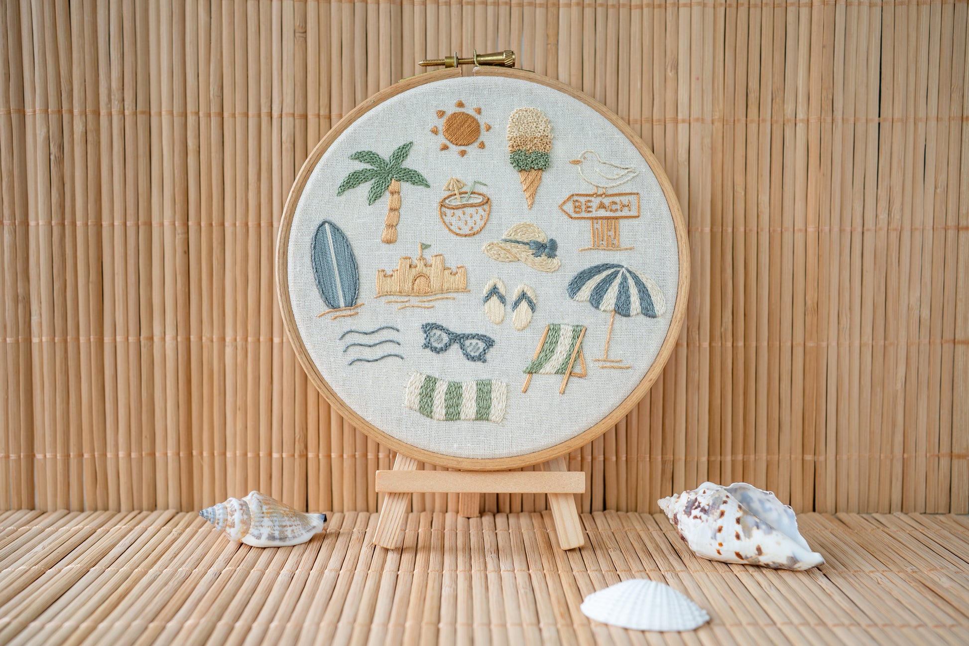 Embroidery hoop with beach-themed designs stood on a mini easel on a wooden surface with shells.