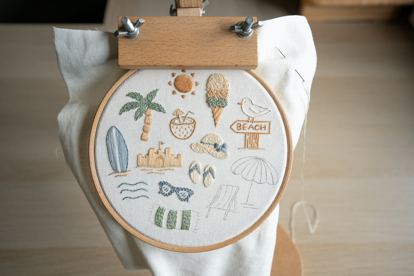 Embroidery hoop with beach-themed designs held by an embroidery hoop stand.