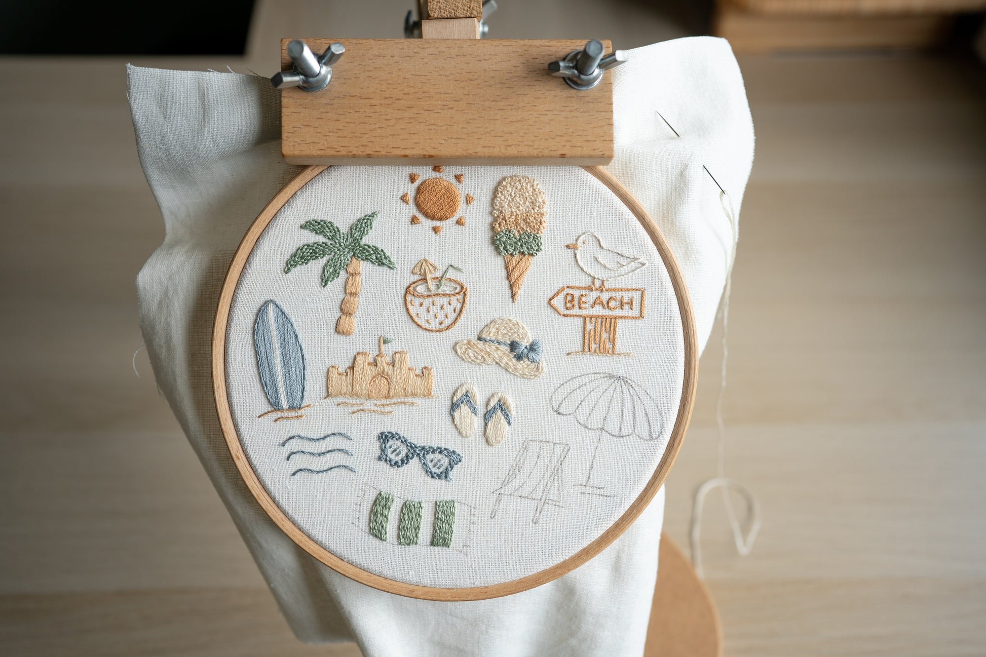 Embroidery hoop with beach-themed designs held by an embroidery hoop stand.