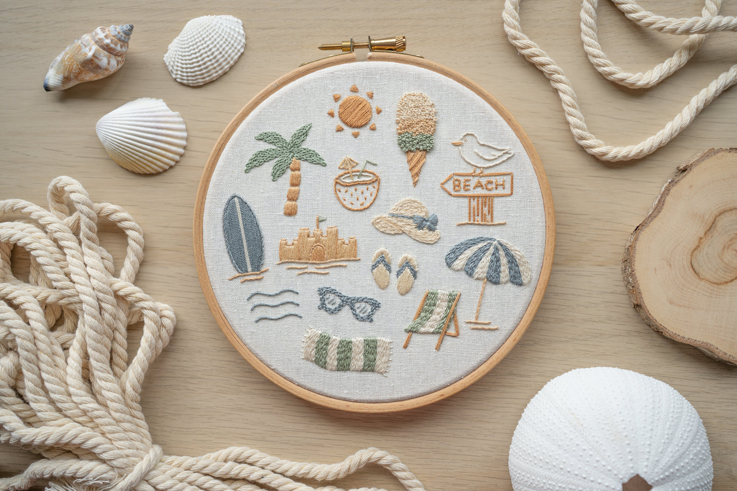 Embroidery hoop with beach-themed designs on a wooden surface with ropes and shells.