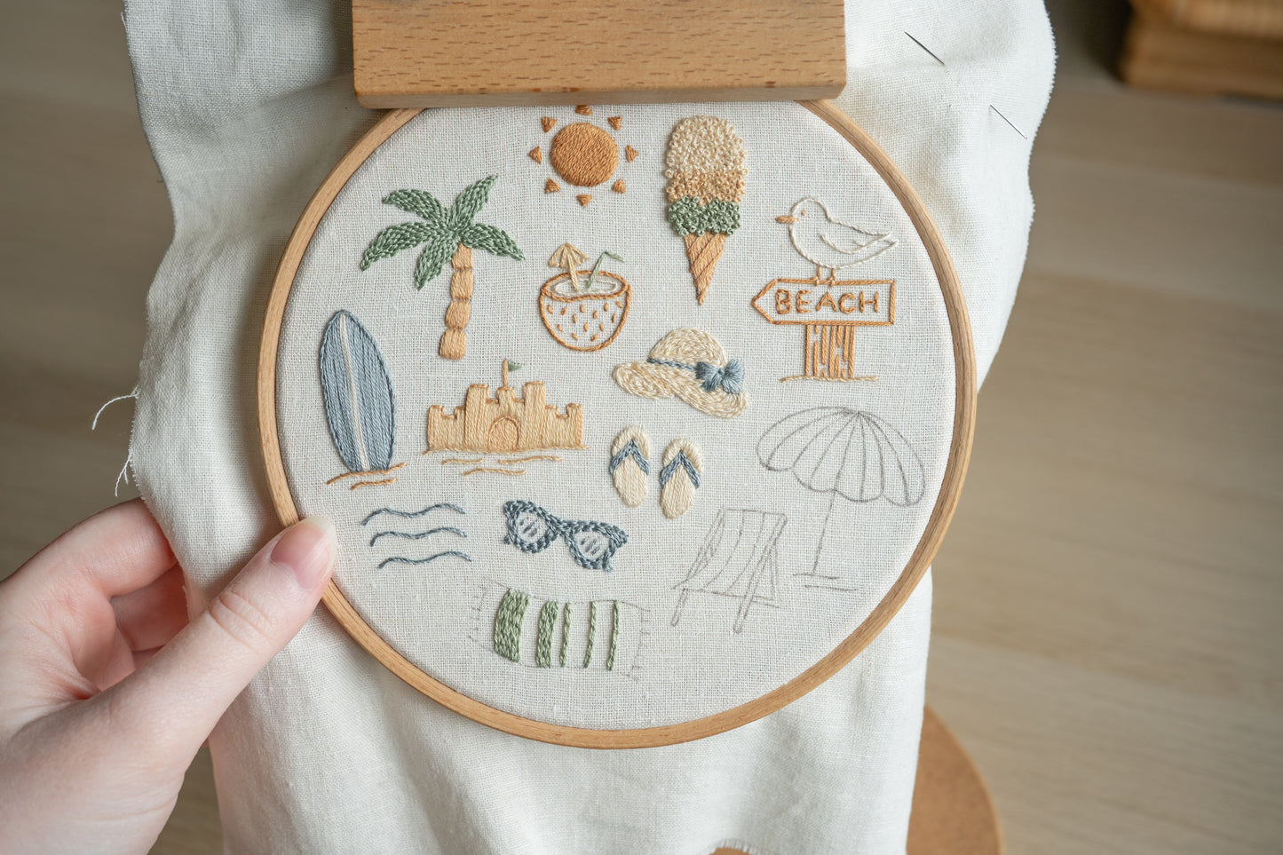 Embroidery hoop with beach-themed designs held by a hand on a wooden surface.