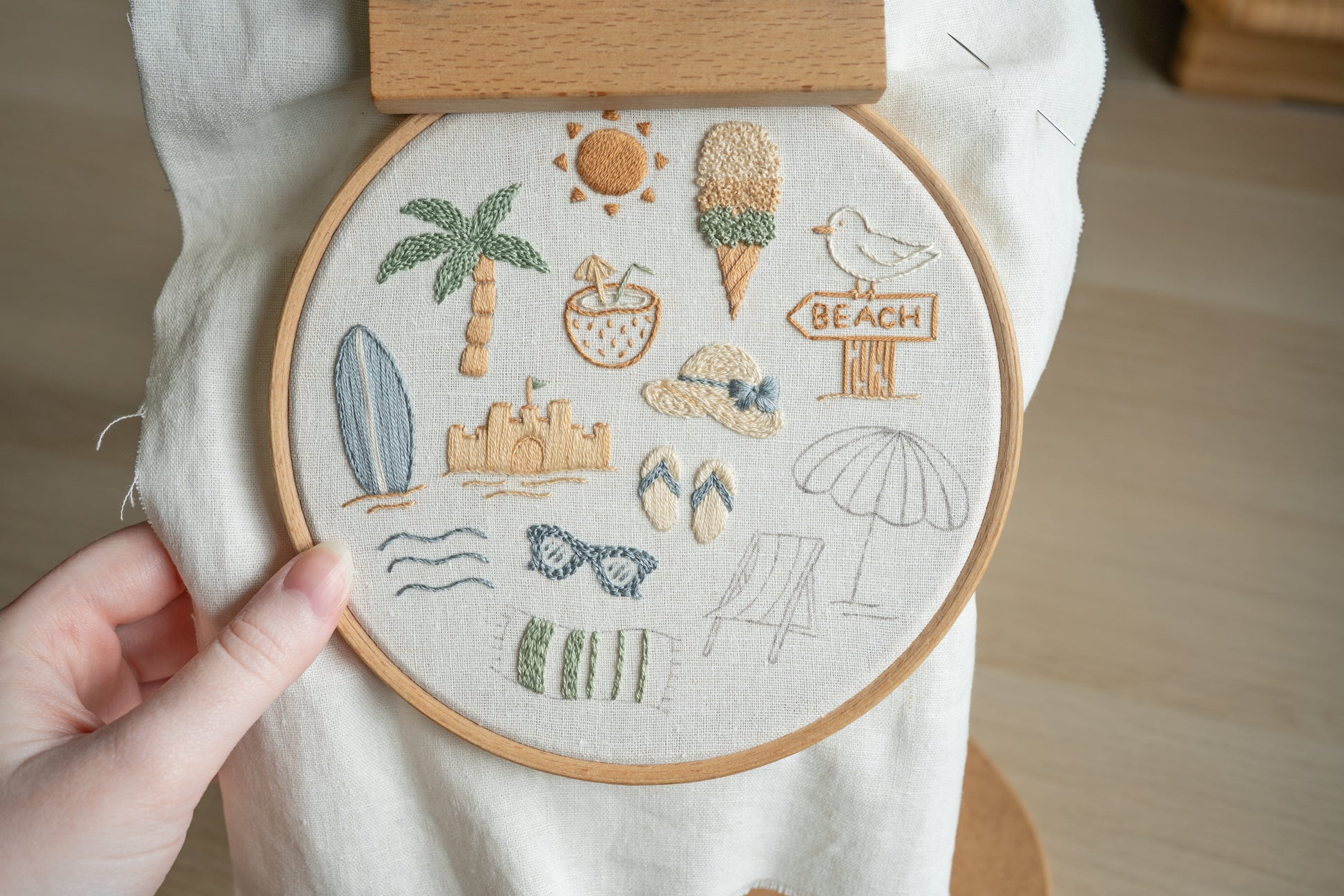 Embroidery hoop with beach-themed designs held by a hand on a wooden surface.