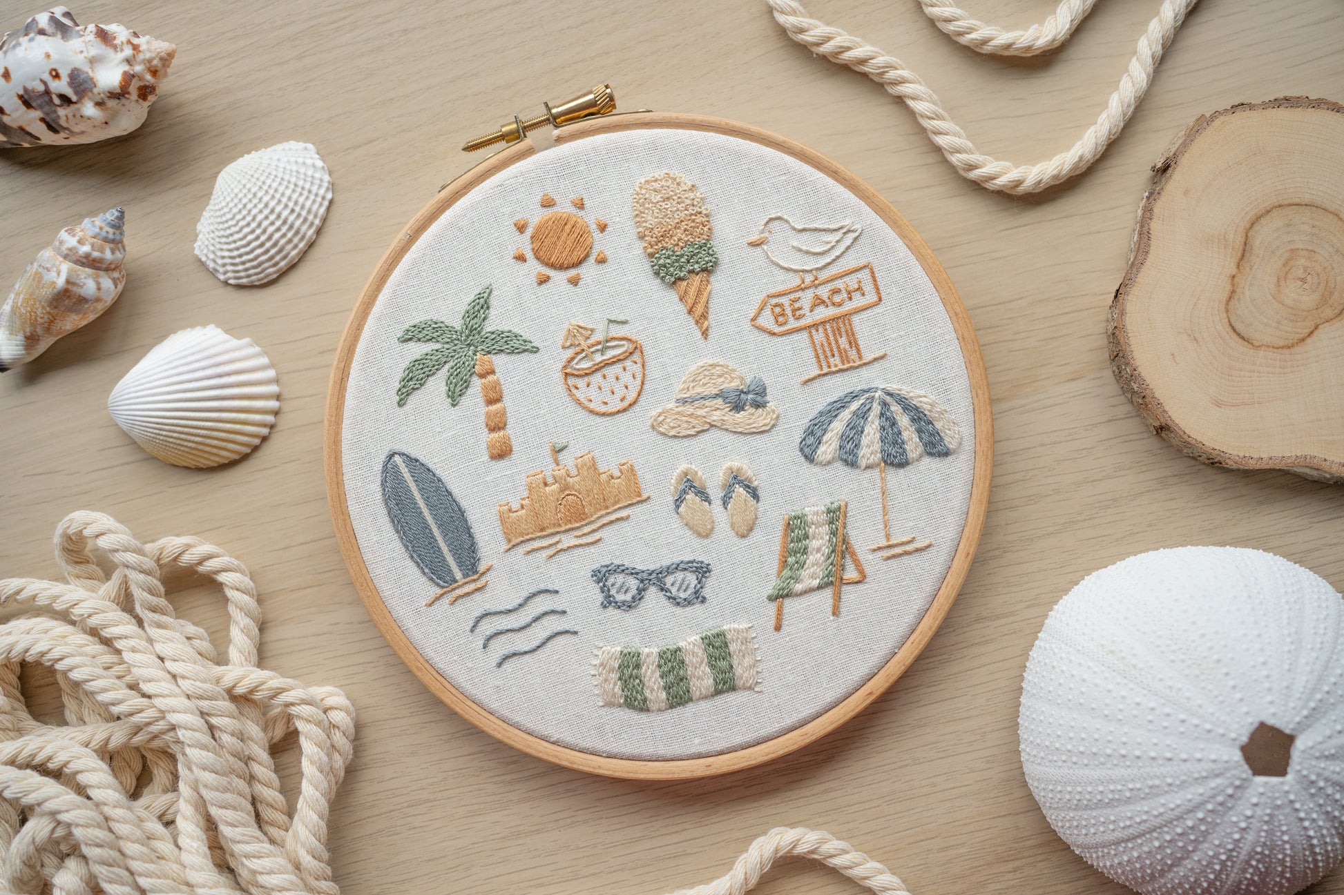 Embroidery hoop with beach-themed designs surrounded by seashells and rope on a wooden surface