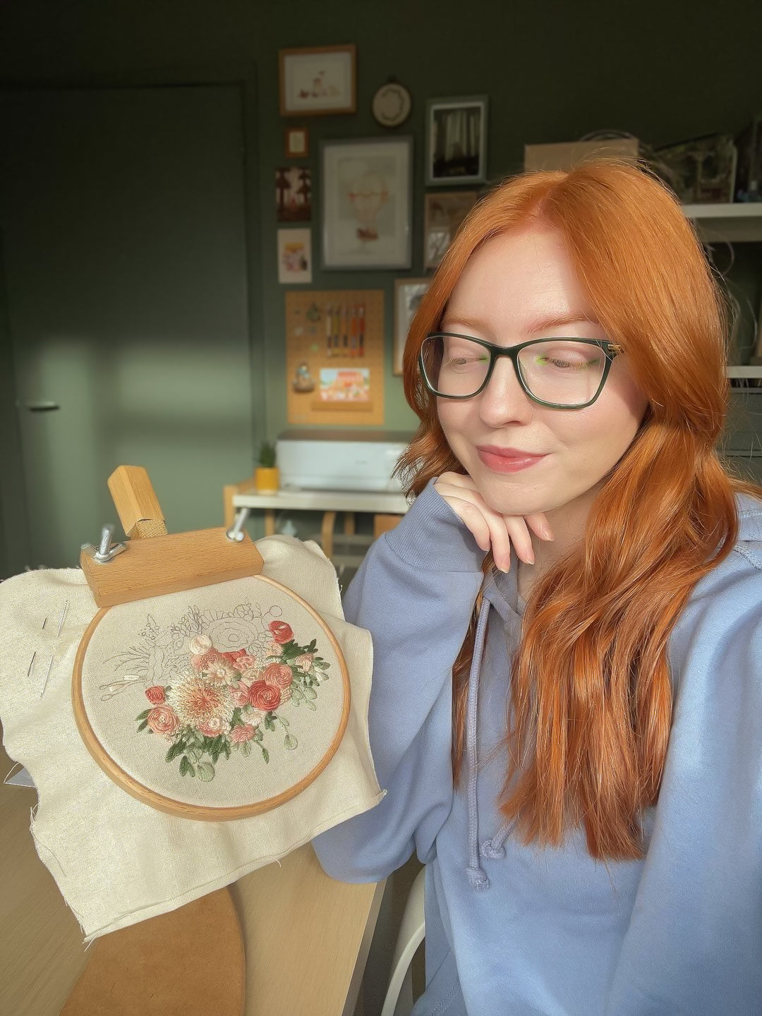 Catherine, a hand embroidery artist, sat at a table with her hand resting under her chin. She's smiling and admiring her half finished embroidered bouquet. Catherine has natural vibrant ginger hair, her skin is pale white, she wears dark green glasses, has vibrant lime green eyeliner in the corners of her eyes, and she's wearing a pale blue hoodie.
