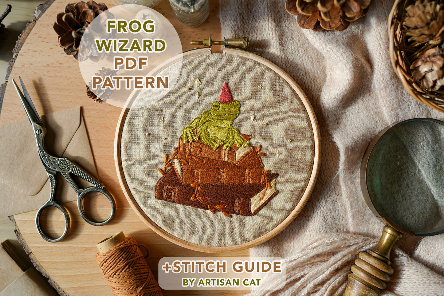 Embroidery hoop with frog wizard design on a wooden board surrounded by nature themed props and sewing tools.