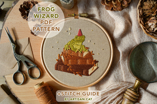 Embroidery hoop with frog wizard design on a wooden board surrounded by nature themed props and sewing tools.