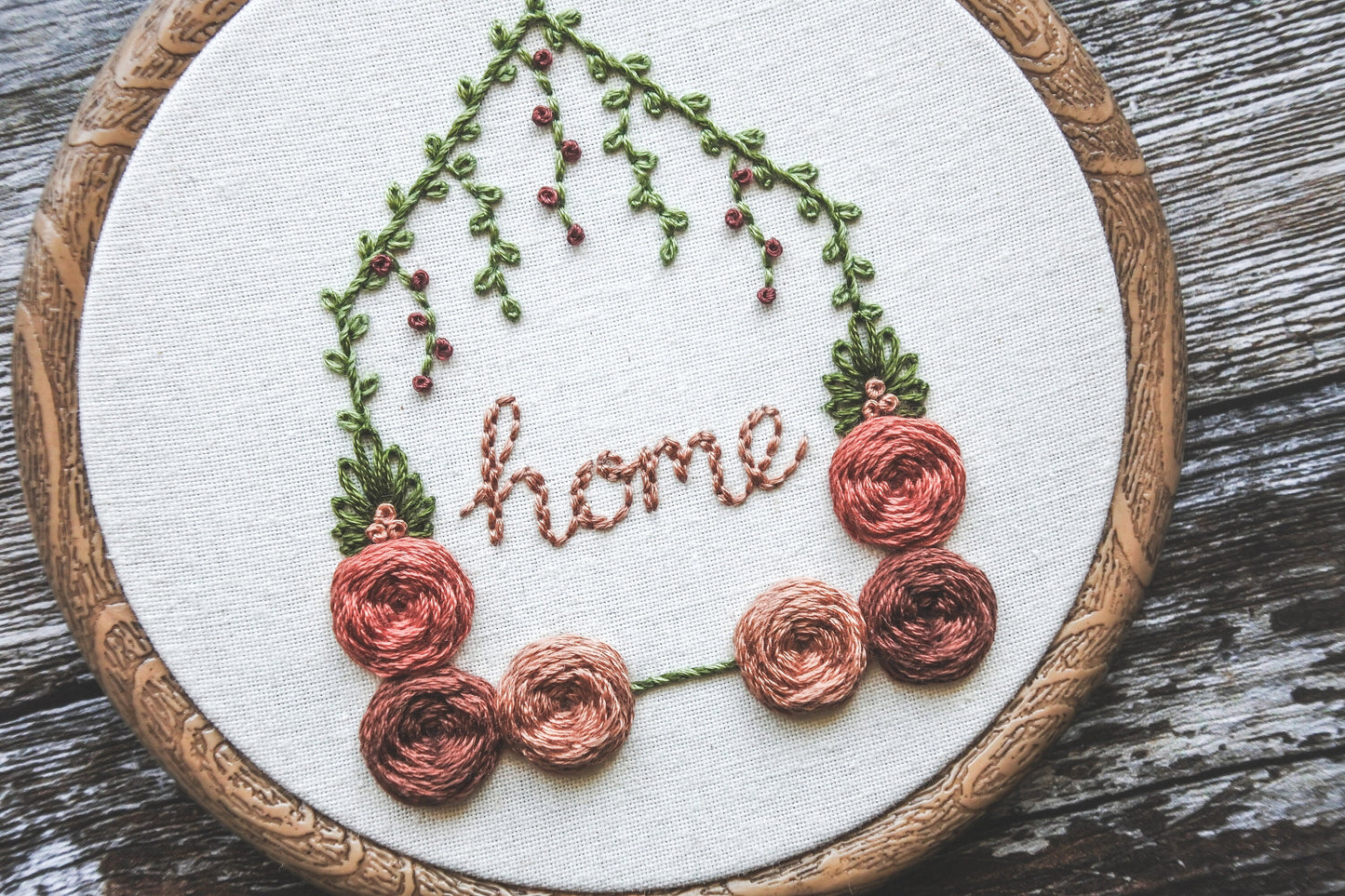 Angled closeup of hand-embroidered floral home hoop with red and peach roses, green foliage, intricate vine details, and the word 'home' in the centre.