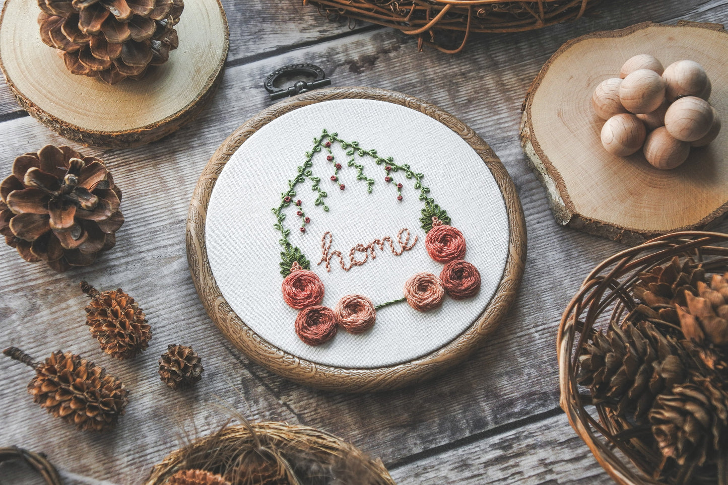 Angled view of hand-embroidered floral home hoop with red and peach colours, green foliage, and the word 'home' in the centre.