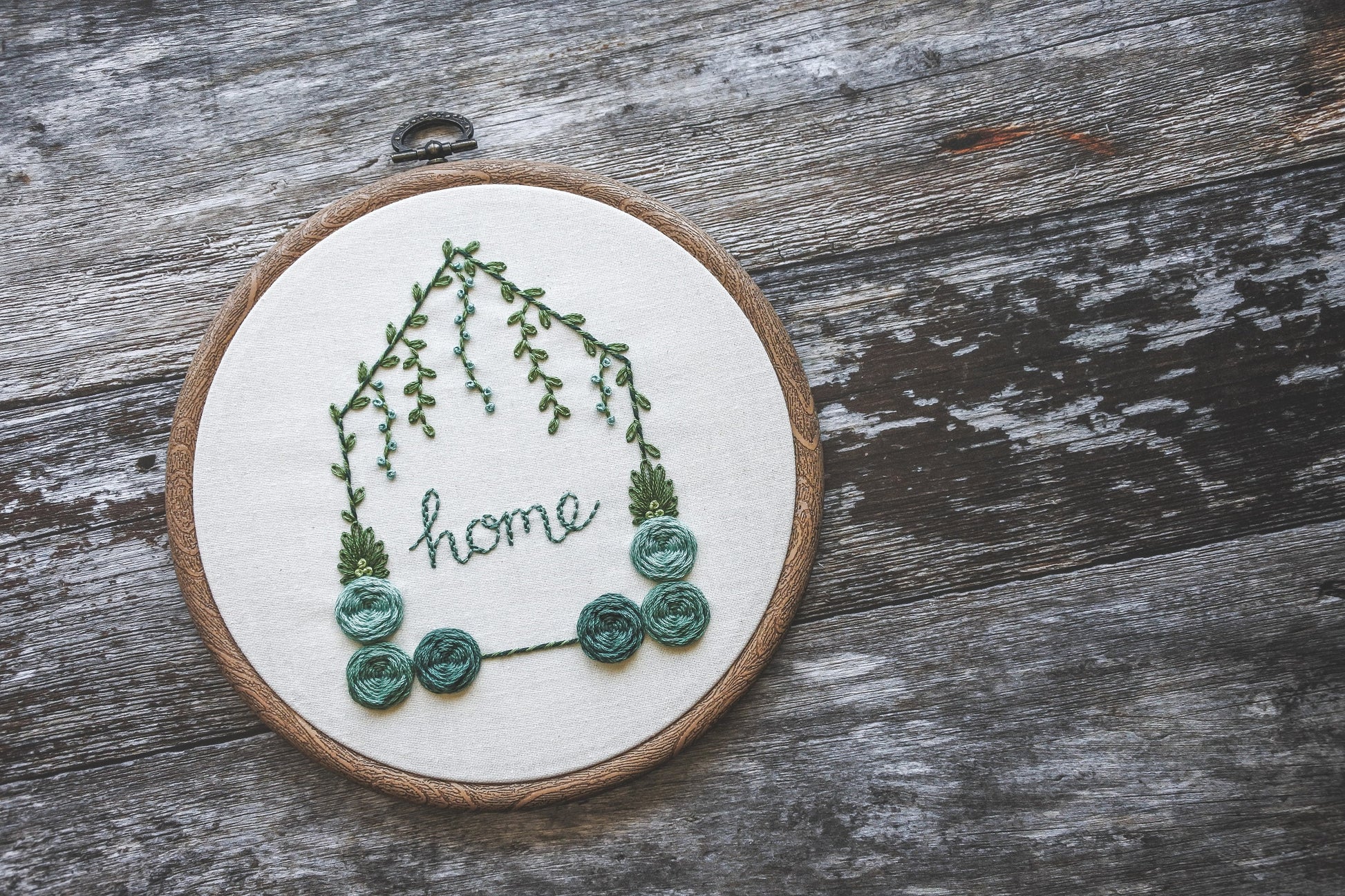 Angled view of hand-embroidered floral home hoop with turquoise colours, green foliage, and the word 'home' in the centre. In a wood effect hoop.
