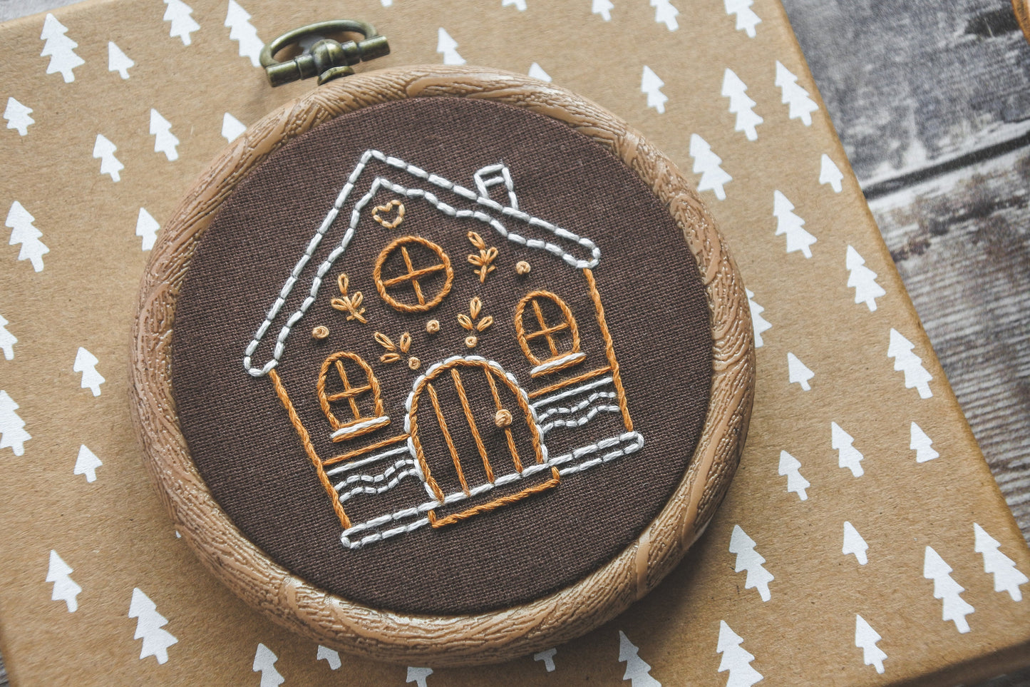 Angled photo of hand-embroidered gingerbread house design in a wood effect hoop, highlighting the stitched details on brown fabric.