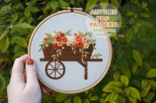 Hand holding an embroidery hoop featuring an autumn wagon design with roses, foliage, and pumpkins, with text reading ‘Autumn PDF pattern + stitch guide by Artisan Cat