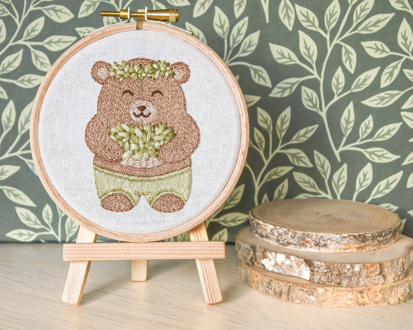 Finished bear embroidery hoop displayed on mini wooden easel with green leaf-patterned background next to mini log slices.
