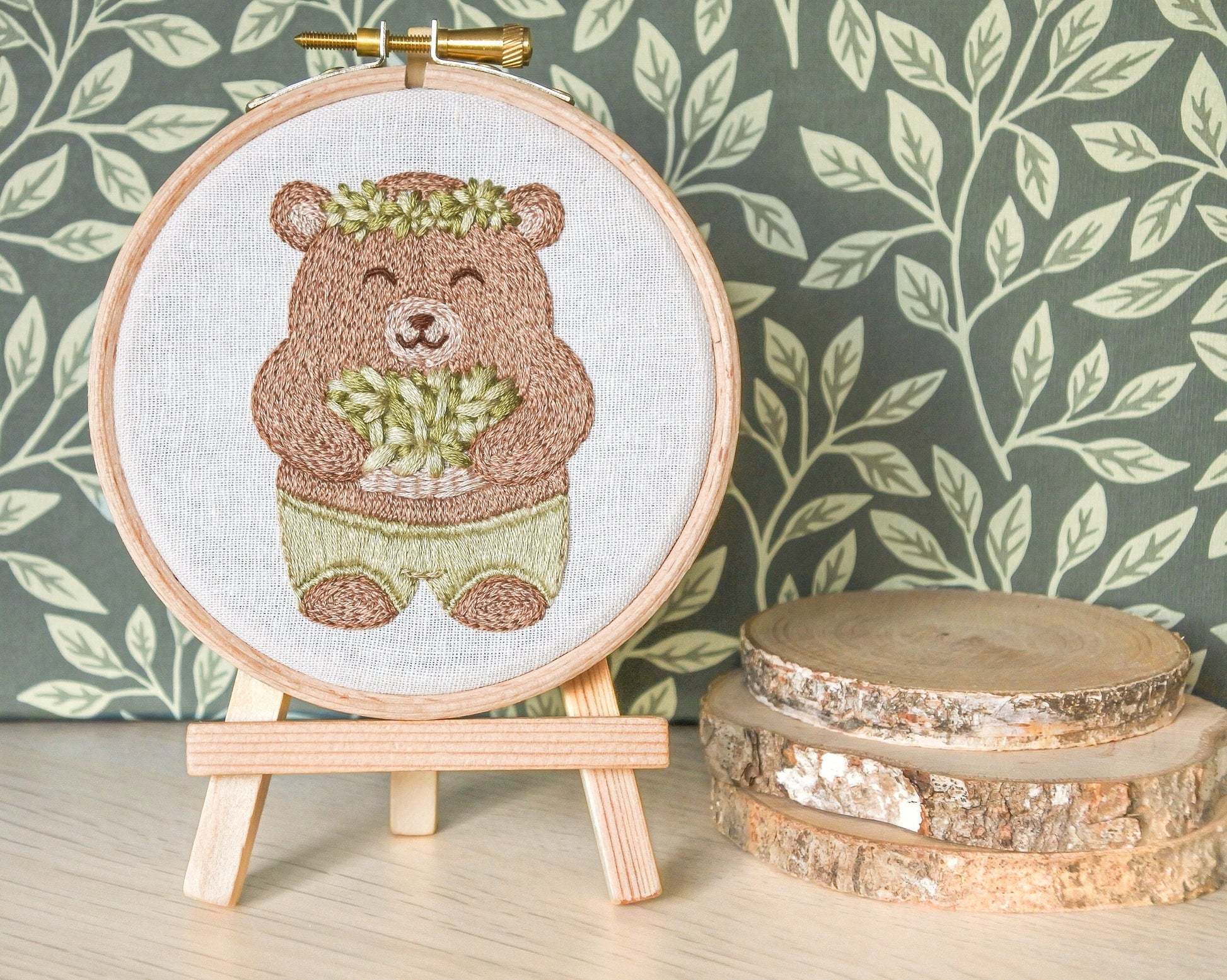 Finished bear embroidery hoop displayed on mini wooden easel with green leaf-patterned background next to mini log slices.