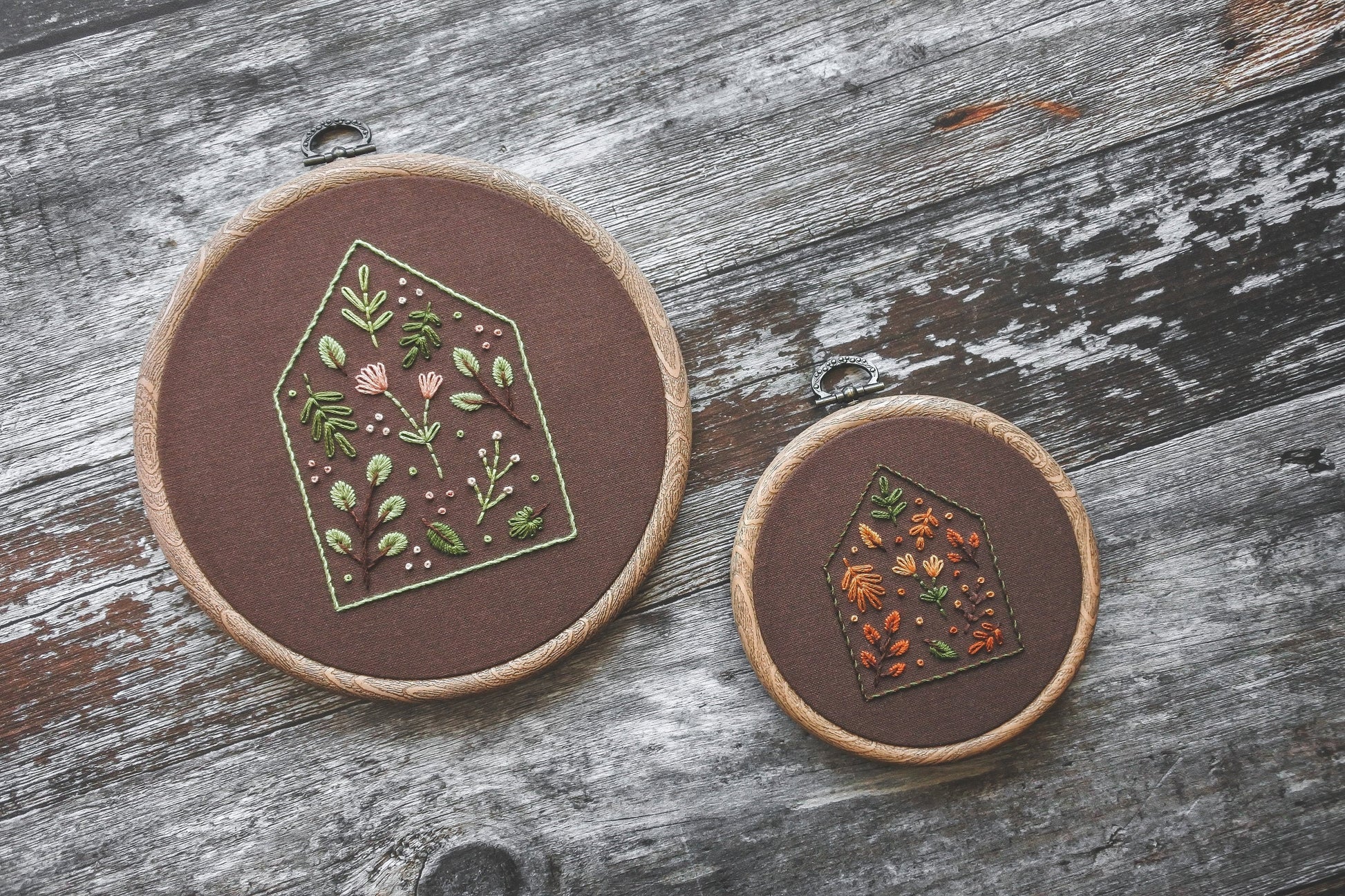 Angled view of two embroidery hoops with house outline designs filled with floral and botanical stitching on dark brown fabric. One hoop has spring colours, the other has warm autumn tones. Both have a wood-effect finish.