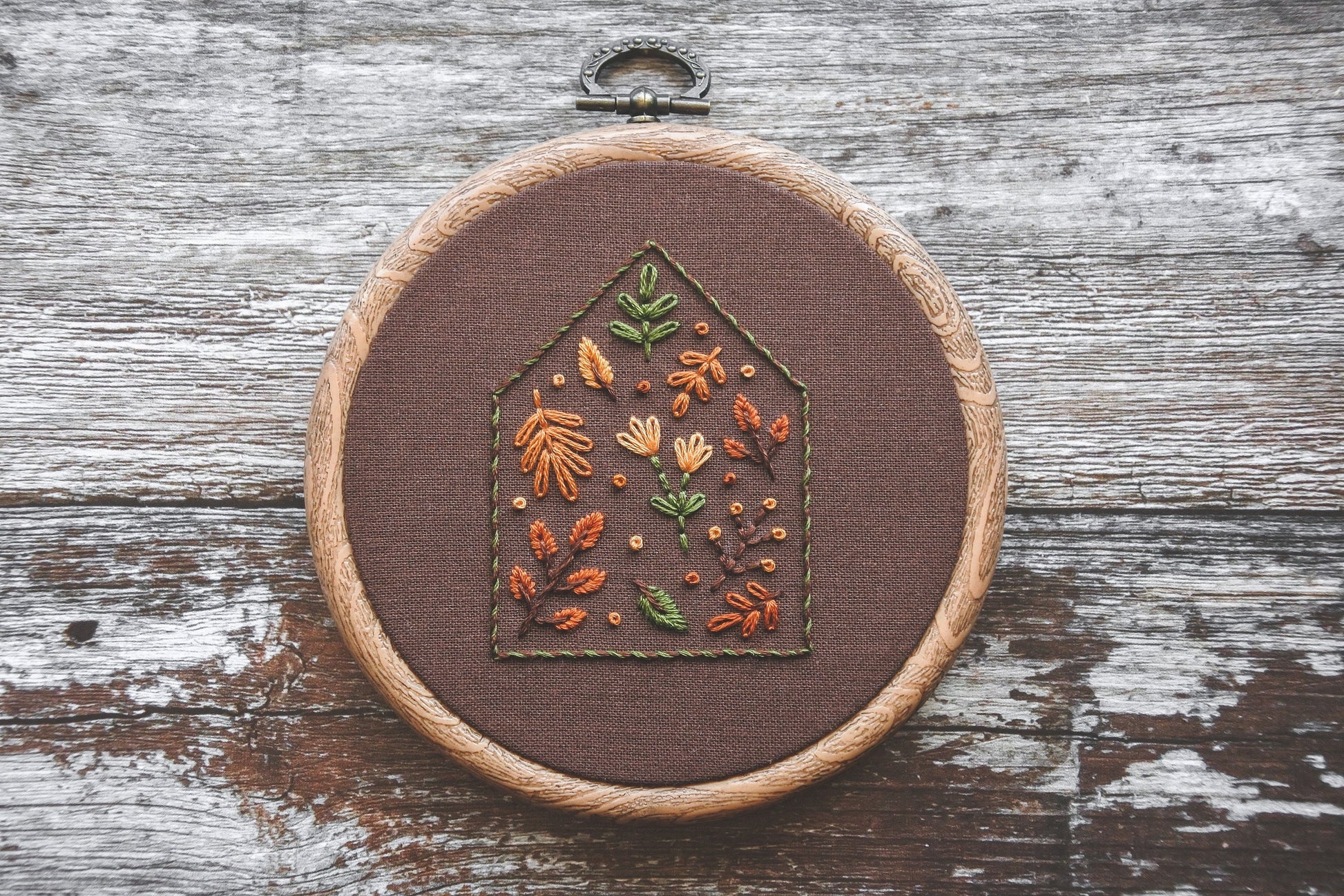 Close-up of a 4-inch embroidery hoop with a house outline filled with autumn-toned floral and botanical stitching on dark brown fabric. The hoop has a wood-effect finish.