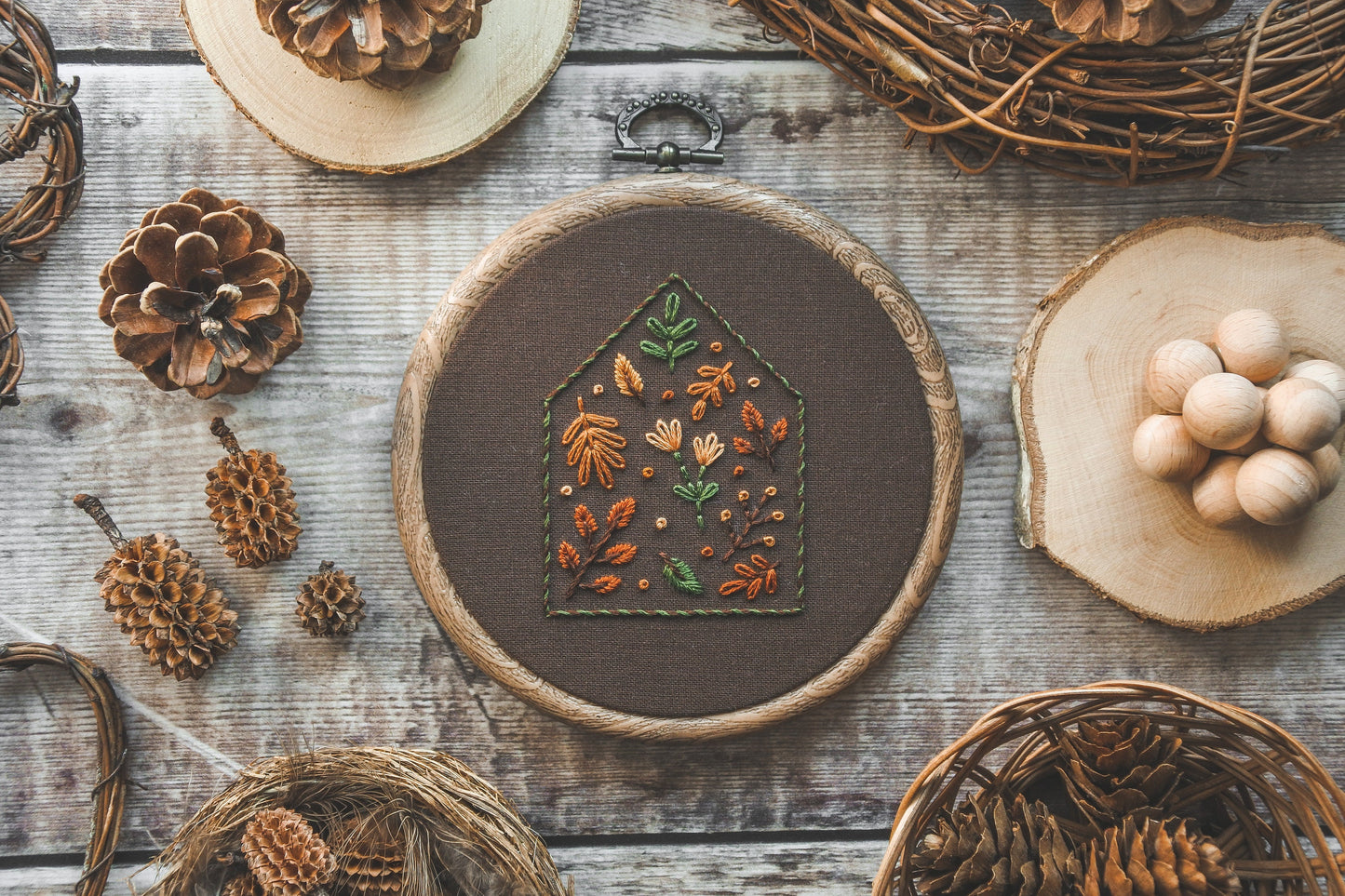 A 4-inch embroidery hoop featuring a house outline filled with warm autumn-coloured floral and botanical stitching on dark brown fabric. The hoop has a wood-effect finish.