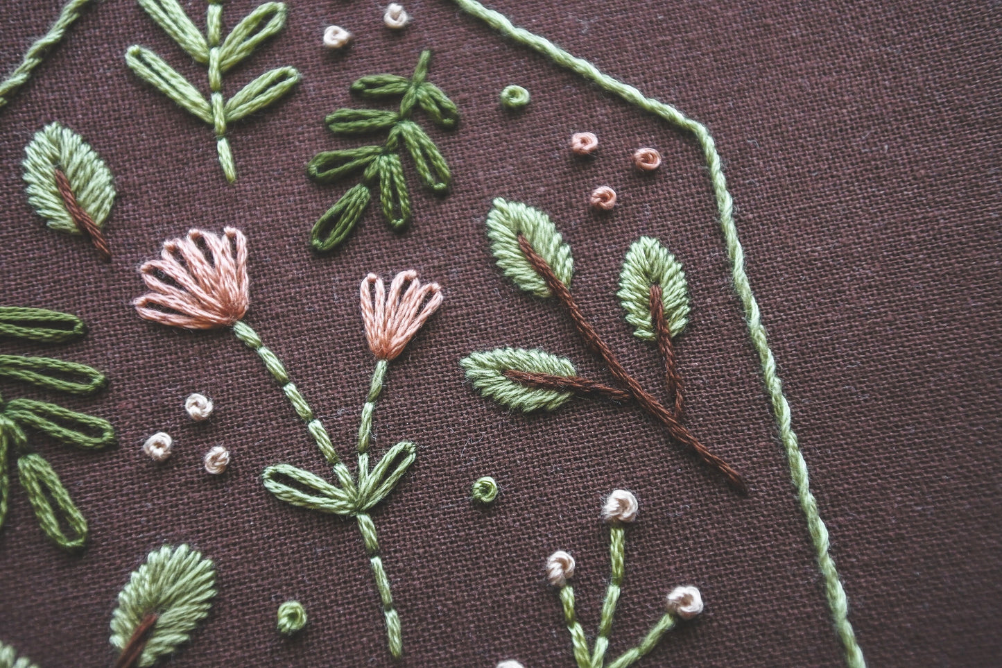 Close-up of an embroidery hoop showcasing the soft spring-toned floral and botanical stitching on dark brown fabric.