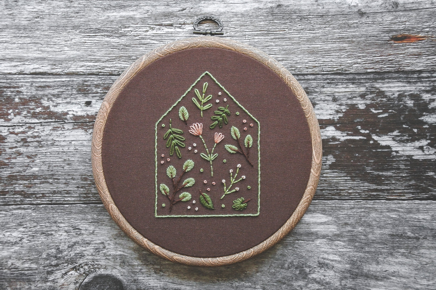 Embroidery hoop featuring a green house outline filled with soft spring-toned floral and botanical stitching on dark brown fabric. The hoop has a wood-effect finish.