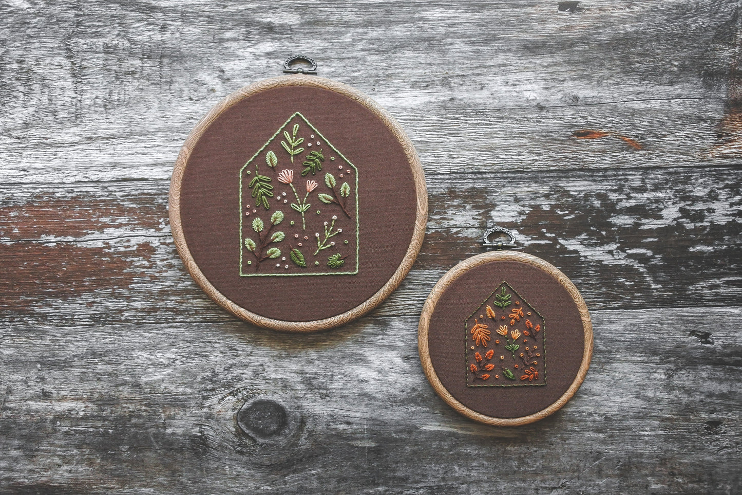 Two embroidery hoops featuring house outlines filled with colourful flowers and foliage, hand stitched on dark brown fabric. One has soft spring tones in a 6-inch hoop, the other features rich autumn colours in a 4-inch hoop. Both hoops have a wood-effect frame.