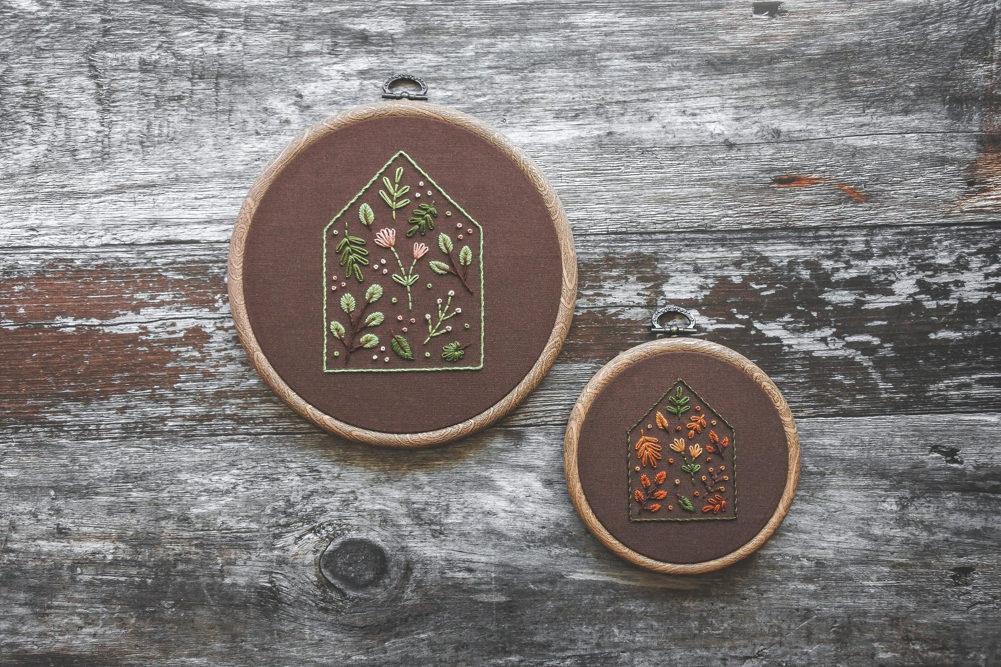 Two embroidery hoops featuring house outlines filled with colourful flowers and foliage, hand stitched on dark brown fabric. One has soft spring tones in a 6-inch hoop, the other features rich autumn colours in a 4-inch hoop. Both hoops have a wood-effect frame.