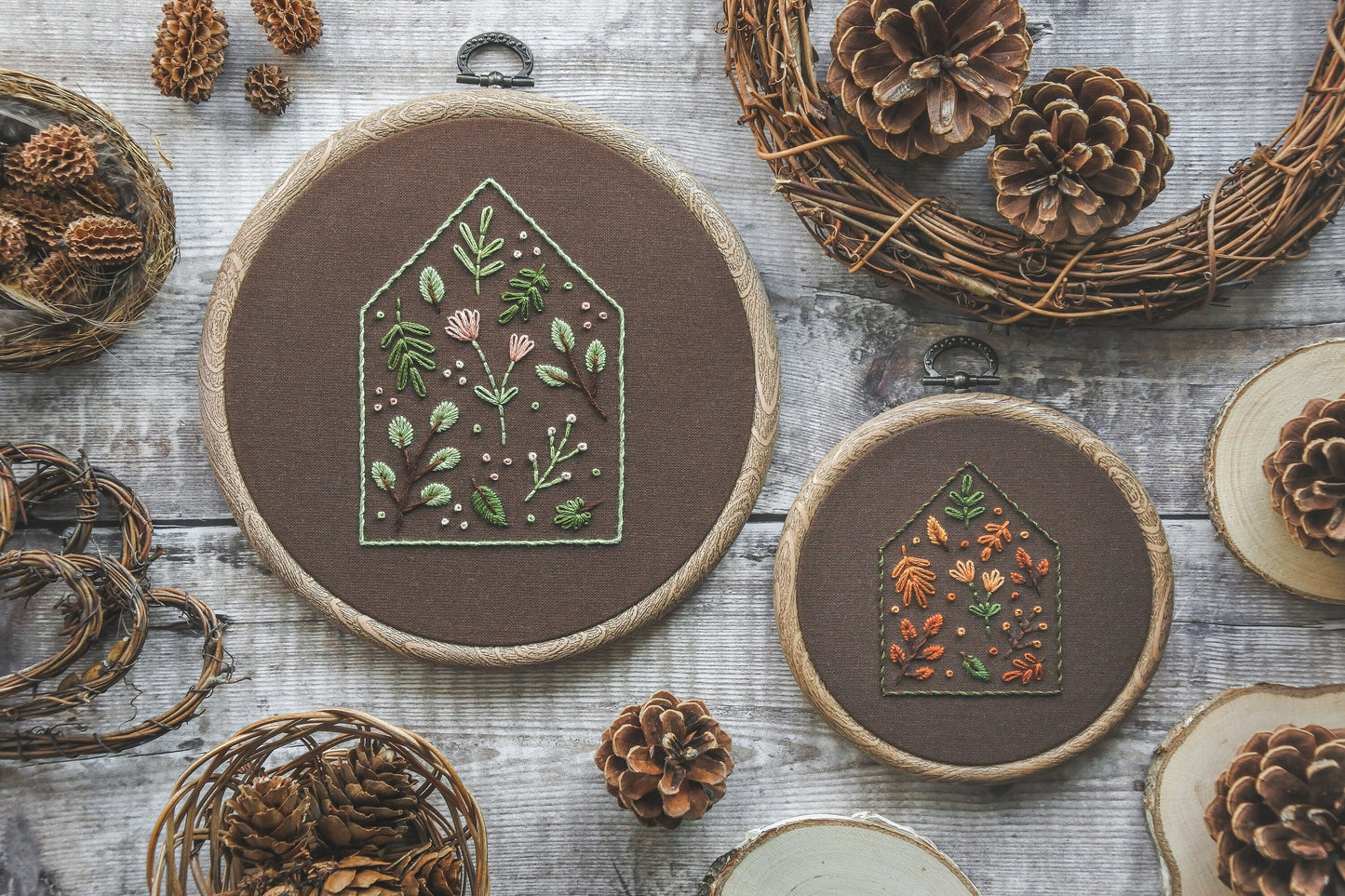 Two embroidery hoops, each featuring a house outline filled with floral and botanical stitching on dark brown fabric. One has a soft spring colour palette in a 6-inch hoop, the other shows warm autumn tones in a 4-inch hoop. Both hoops have a wood-effect finish.