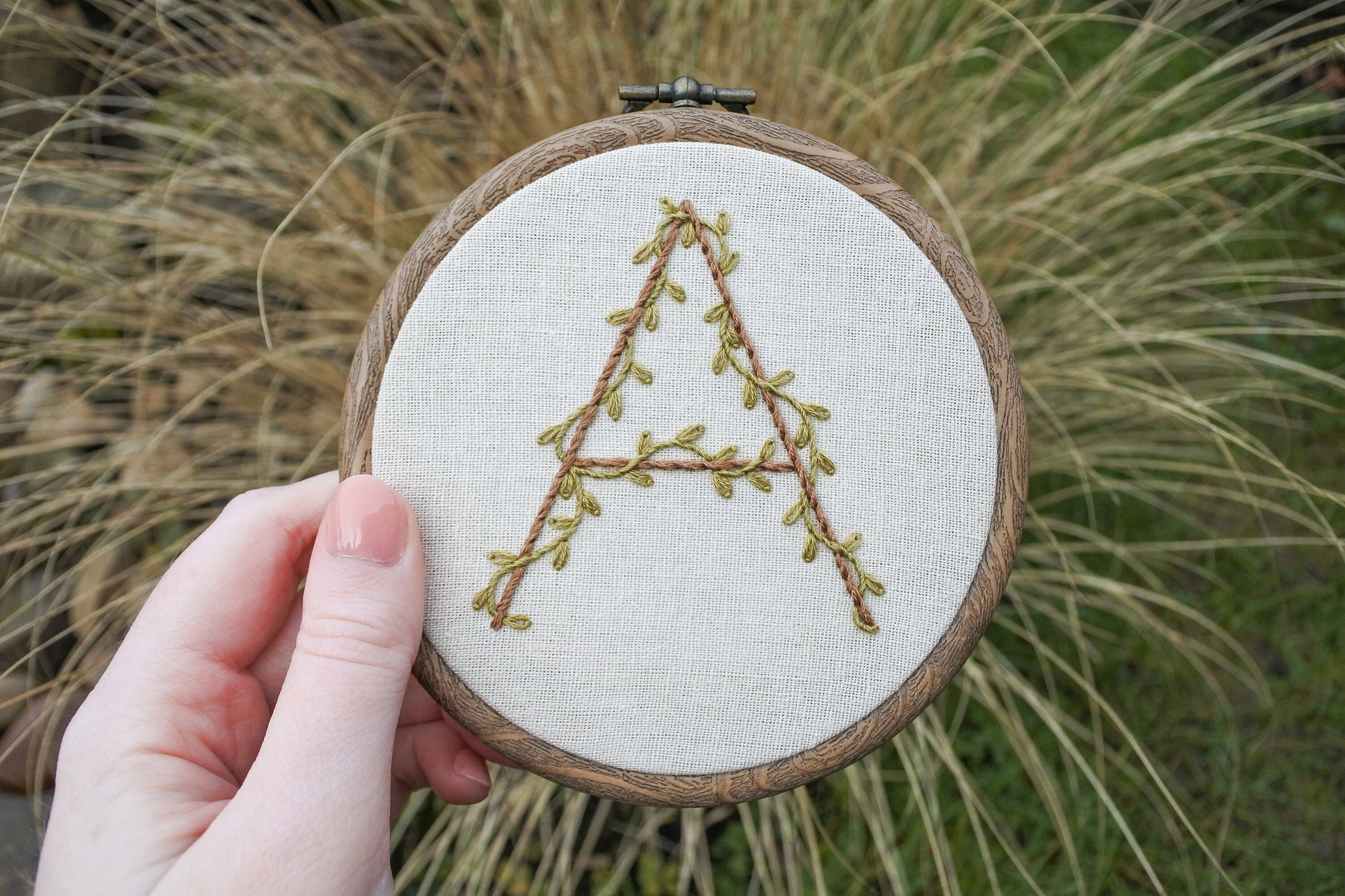 Hand holding the botanical letter 'A' embroidery hoop, stitched with olive green vines and light brown thread on neutral fabric.