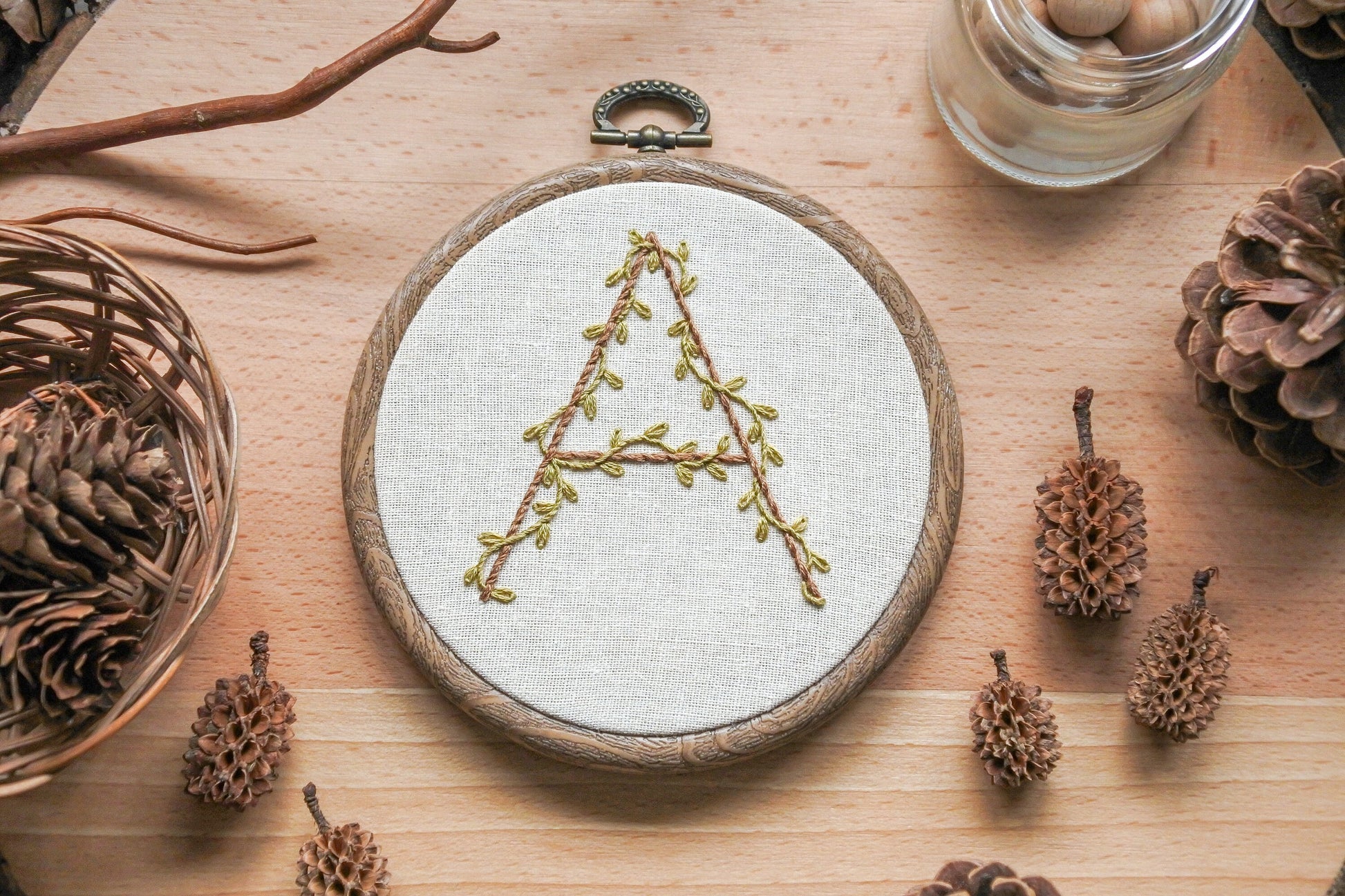 Hand-embroidered botanical letter 'A' hoop with olive green vines and light brown embroidery, set in a 4-inch wood-effect hoop.