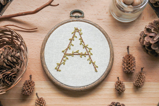 Hand-embroidered botanical letter 'A' hoop with olive green vines and light brown embroidery, set in a 4-inch wood-effect hoop.