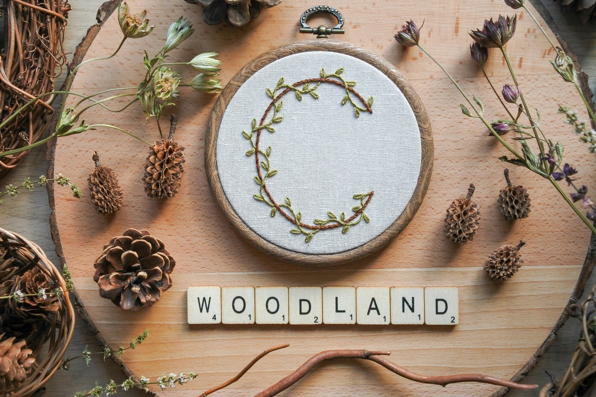 Botanical letter C embroidery hoop with green vines and brown stitched letter, displayed with wooden Scrabble tiles spelling 'woodland'