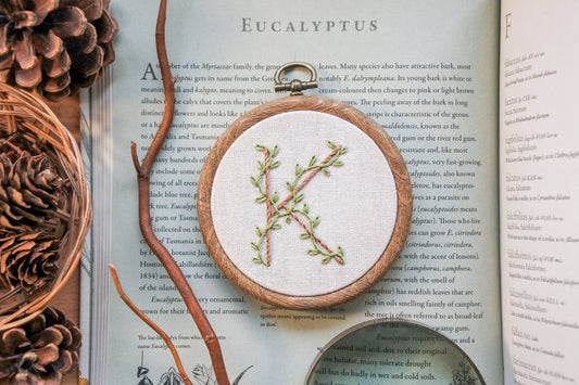 Botanical letter K embroidery hoop with eucalyptus green vines and pale brown stitched letter on neutral fabric, framed in a 3-inch wood-effect hoop