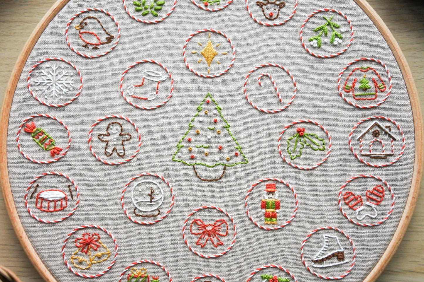 Hand embroidered Christmas advent calendar design, showing festive elements stitched in red, green, gold and white on light beige fabric.