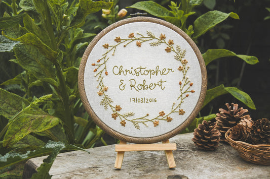 Personalised autumn embroidery hoop with the names Christopher and Robert, and date underneath, displayed on a mini wooden easel outside with greenery and pinecones
