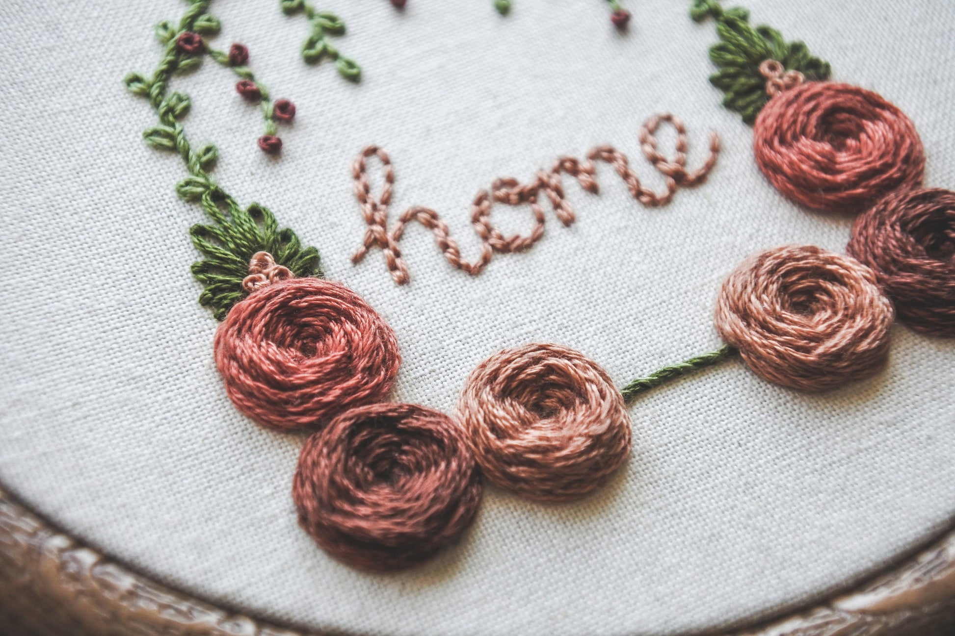 Closeup of hand-embroidered muted red and peach roses with green foliage on a floral home embroidery hoop.