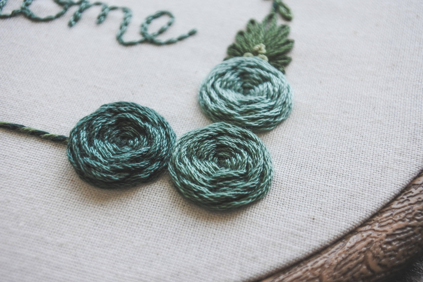 Closeup of hand-embroidered turquoise roses with green foliage on a floral home embroidery hoop.