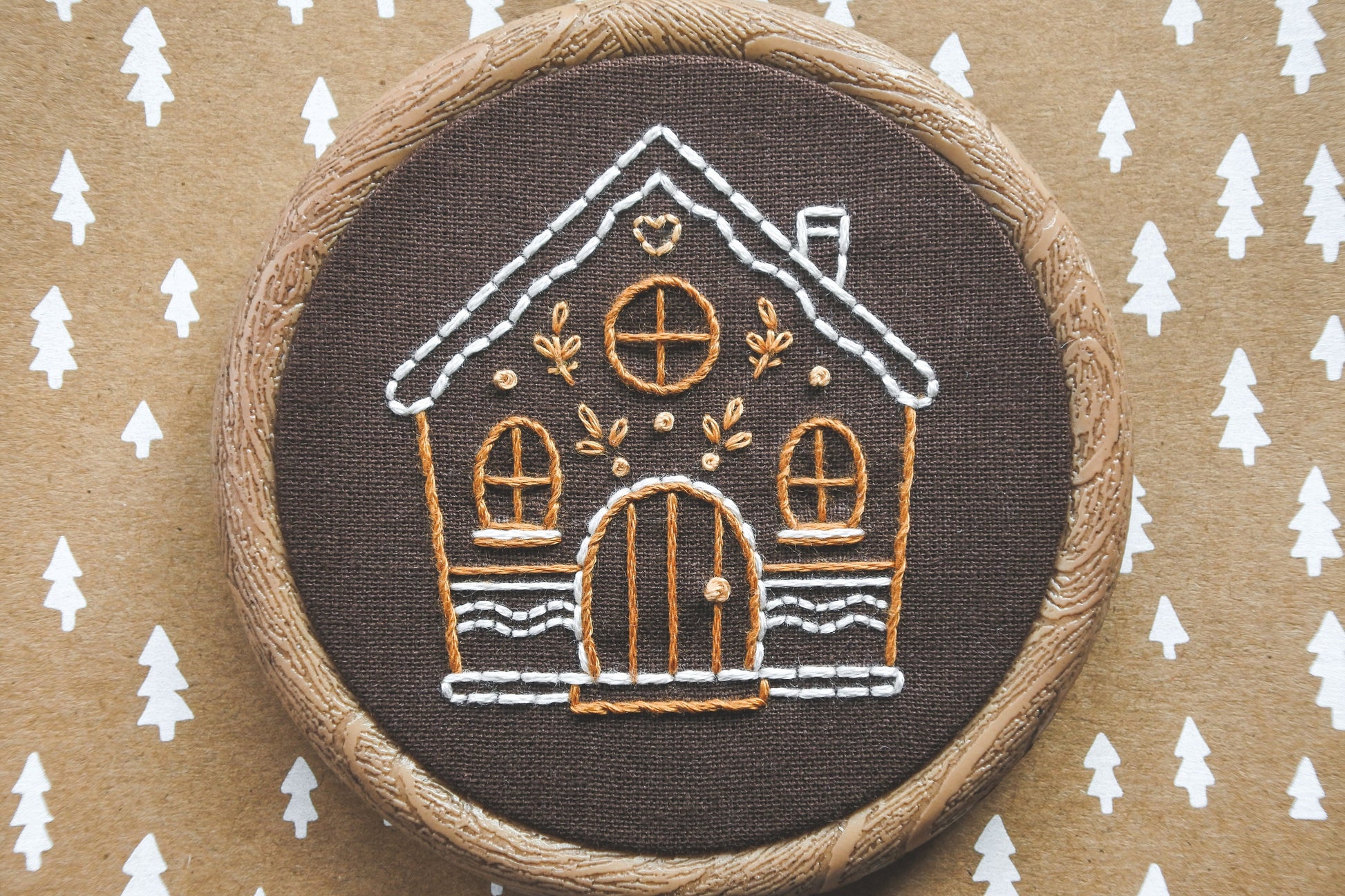 Close-up of hand-embroidered gingerbread house design in hoop, focusing on stitching details on brown fabric.