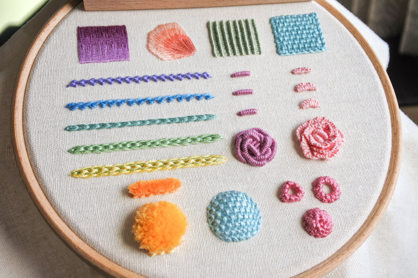 Angled view of a colourful finished hand embroidery stitch sampler featuring 12 stitches on fabric in a wooden hoop