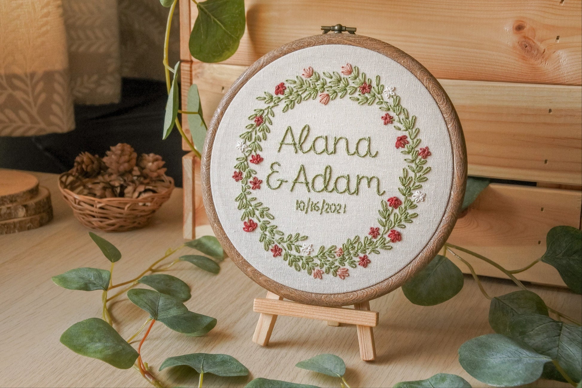 Custom summer wreath embroidery hoop with “Alana and Adam” and their wedding date, stitched with muted red, pink and white flowers, displayed on easel surrounded by a eucalyptus leaf garland