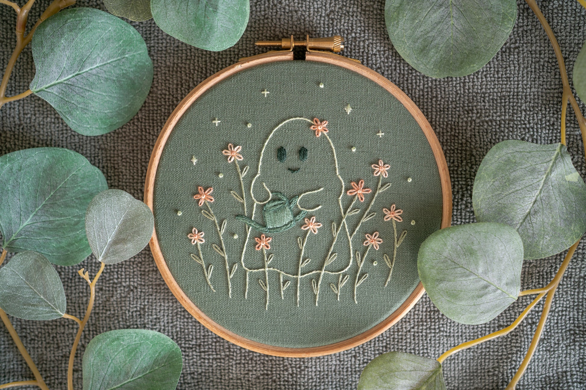 hand embroidered cute ghost watering flowers. The embroidery hoop is surrounded by artificial eucalyptus foliage.